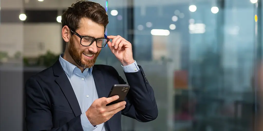 Mann checkt sein Handy im gläsernen Büro, fass mit Hand an die Brille.
